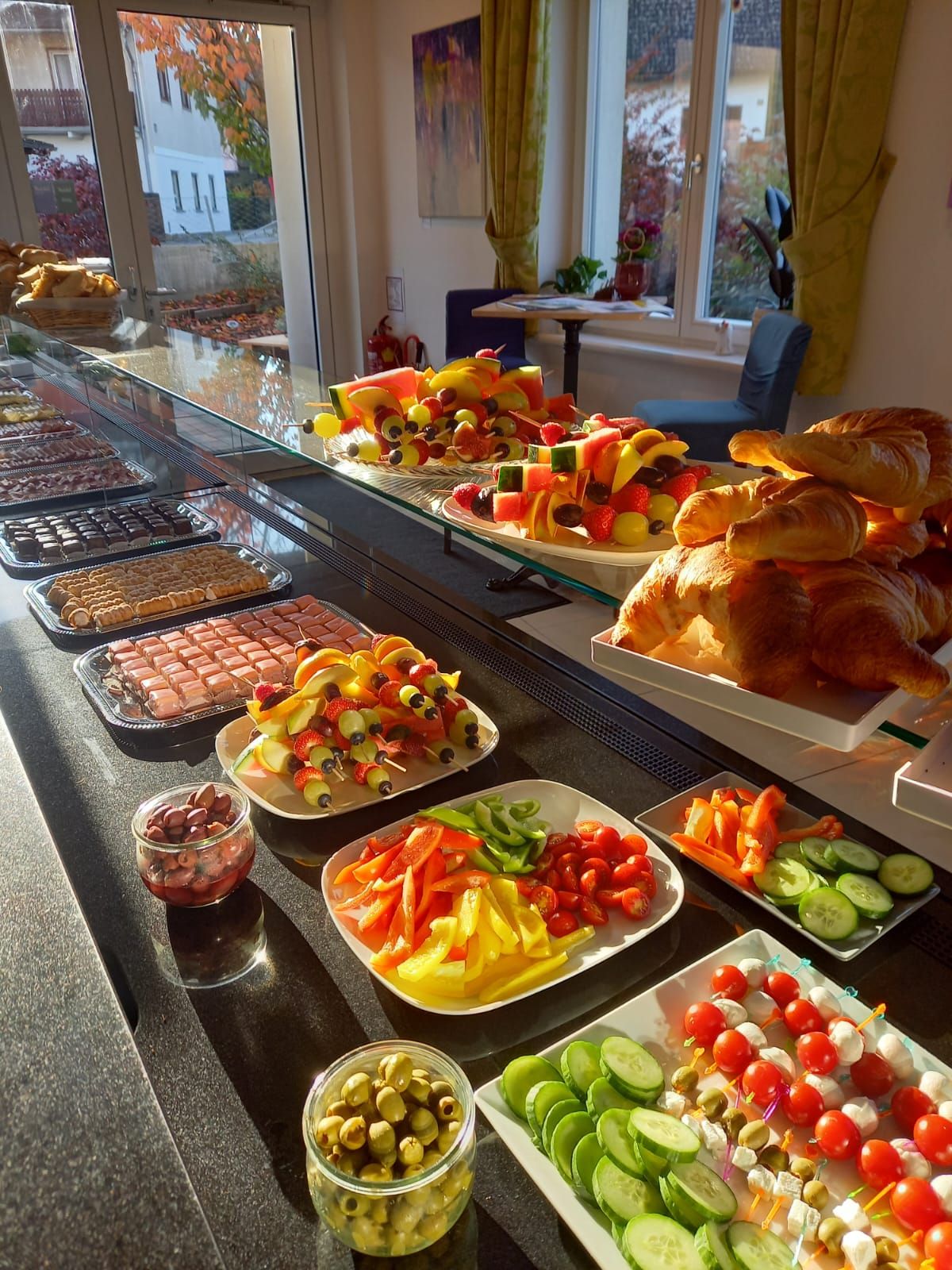 A buffet with croissants, fruit skewers, vegetables and olives in a sun-drenched room.