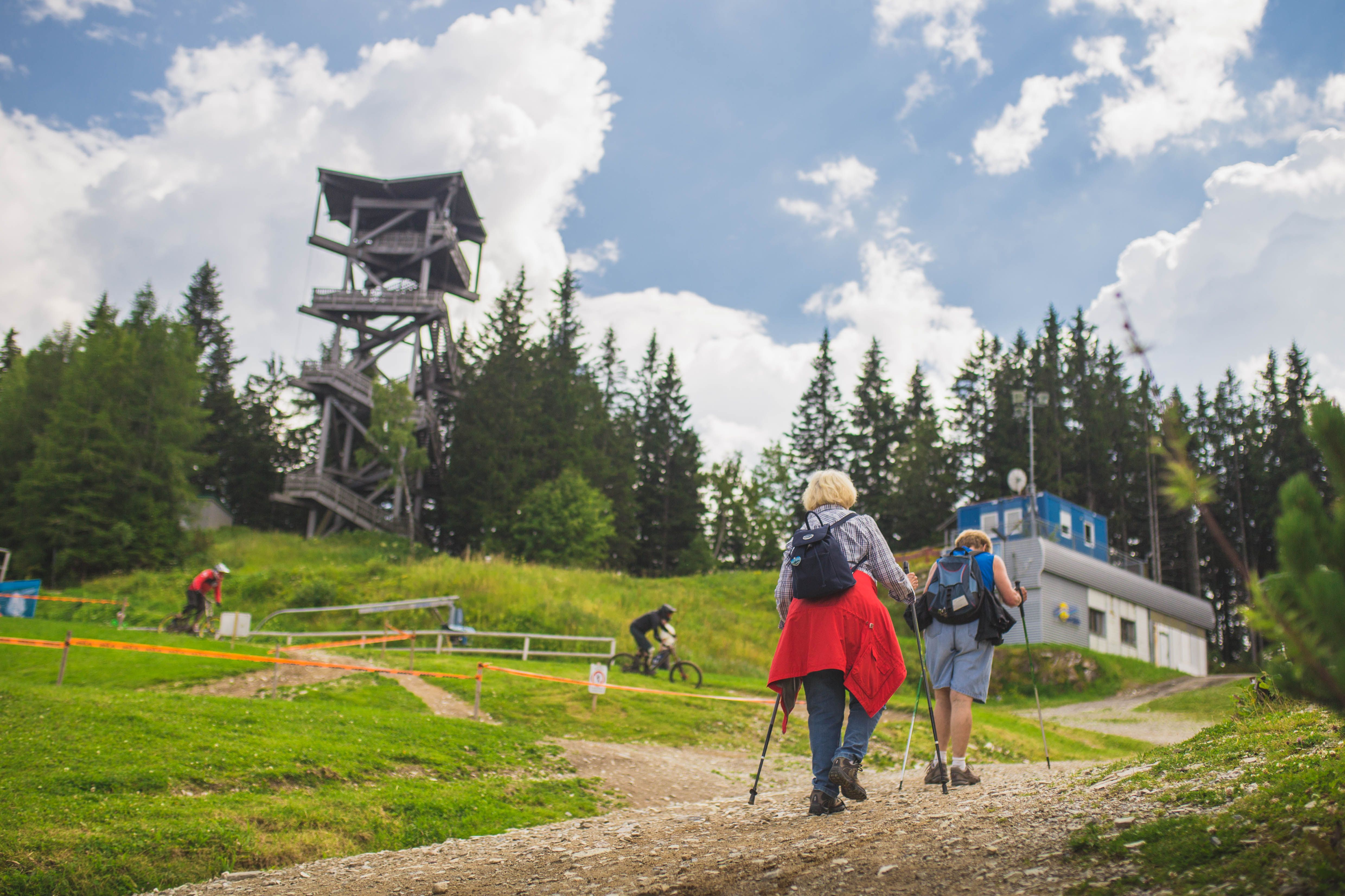 Two hikers walk uphill on a path, past an observation tower and a building, surrounded by trees and a clear sky.