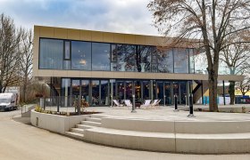 Modern two-storey building with glass fa&ccedil;ade and terrace, surrounded by trees.