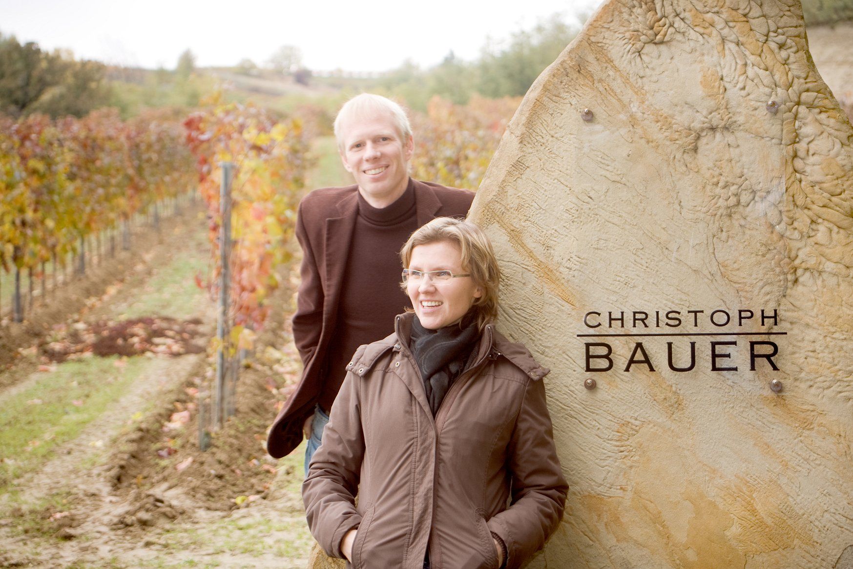 Two people stand smiling in front of a vineyard next to a stone with the inscription 'Christoph Bauer'.