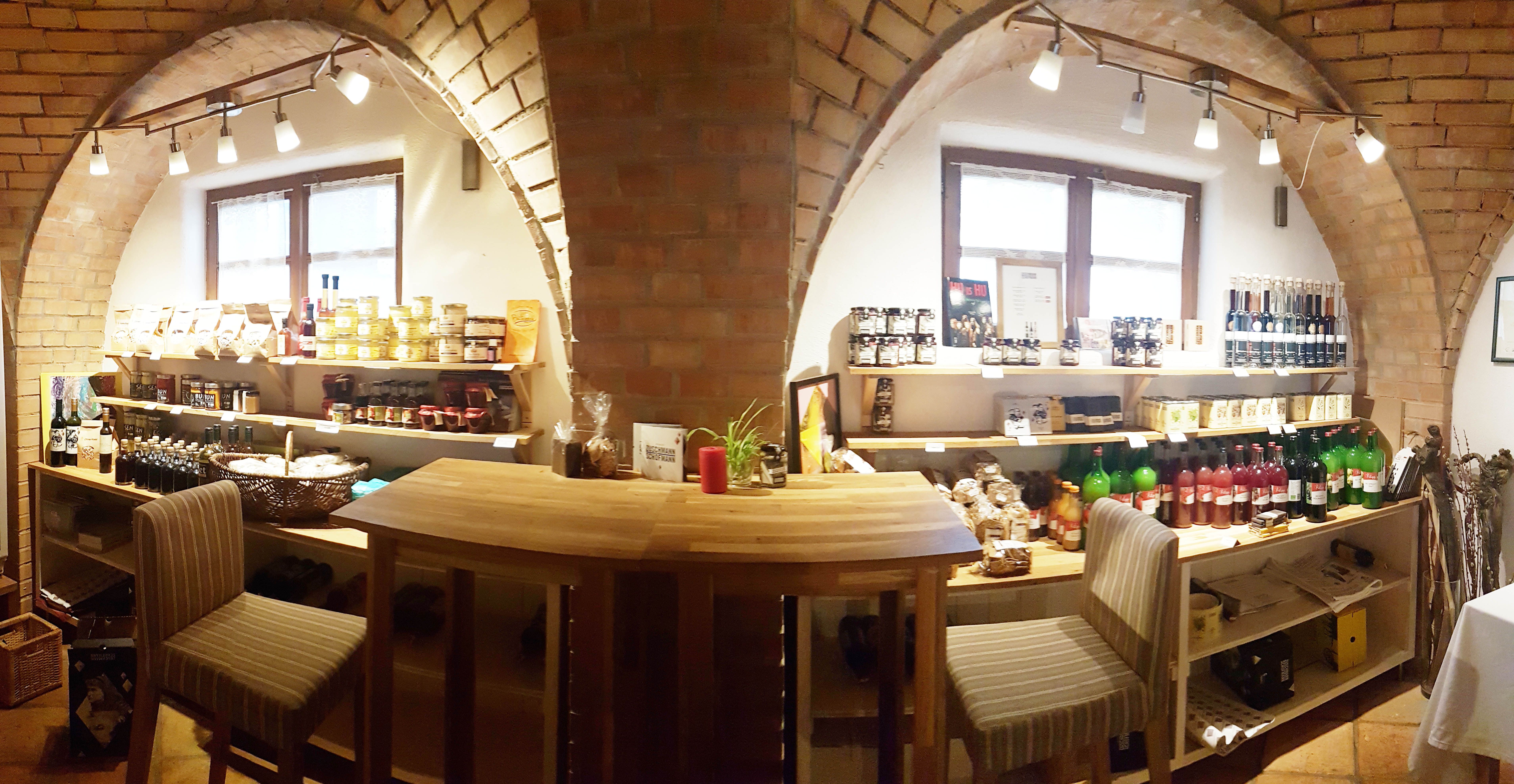 Interior view of a small store with shelves full of food and drinks, wooden table and chairs.