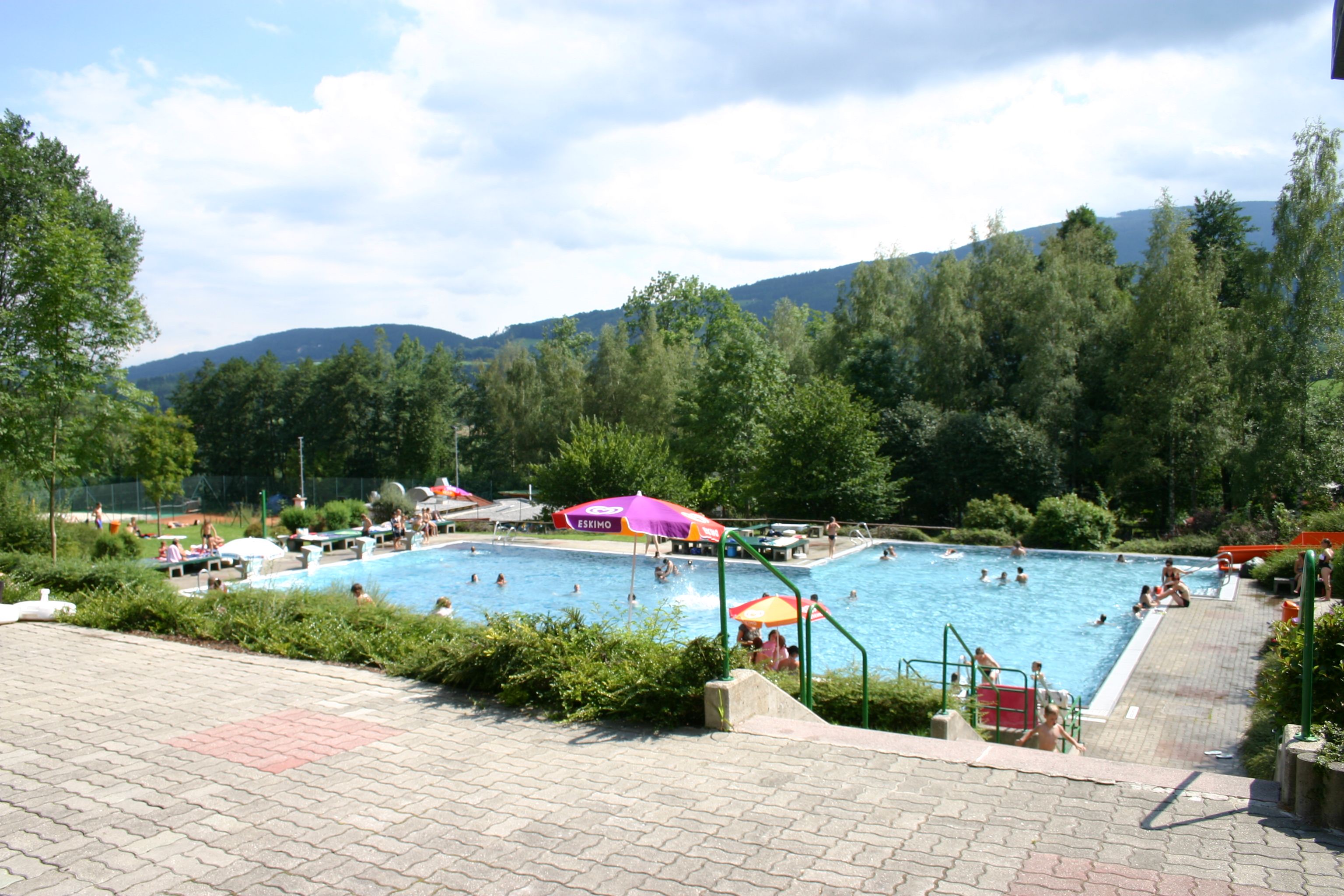Outdoor pool with floats and parasols, surrounded by trees and hills.