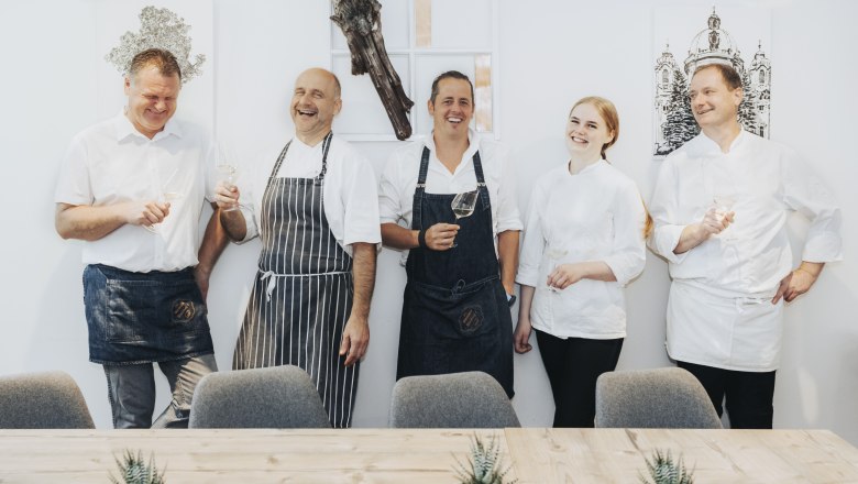 Five people dressed as chefs are laughing and holding glasses of wine.