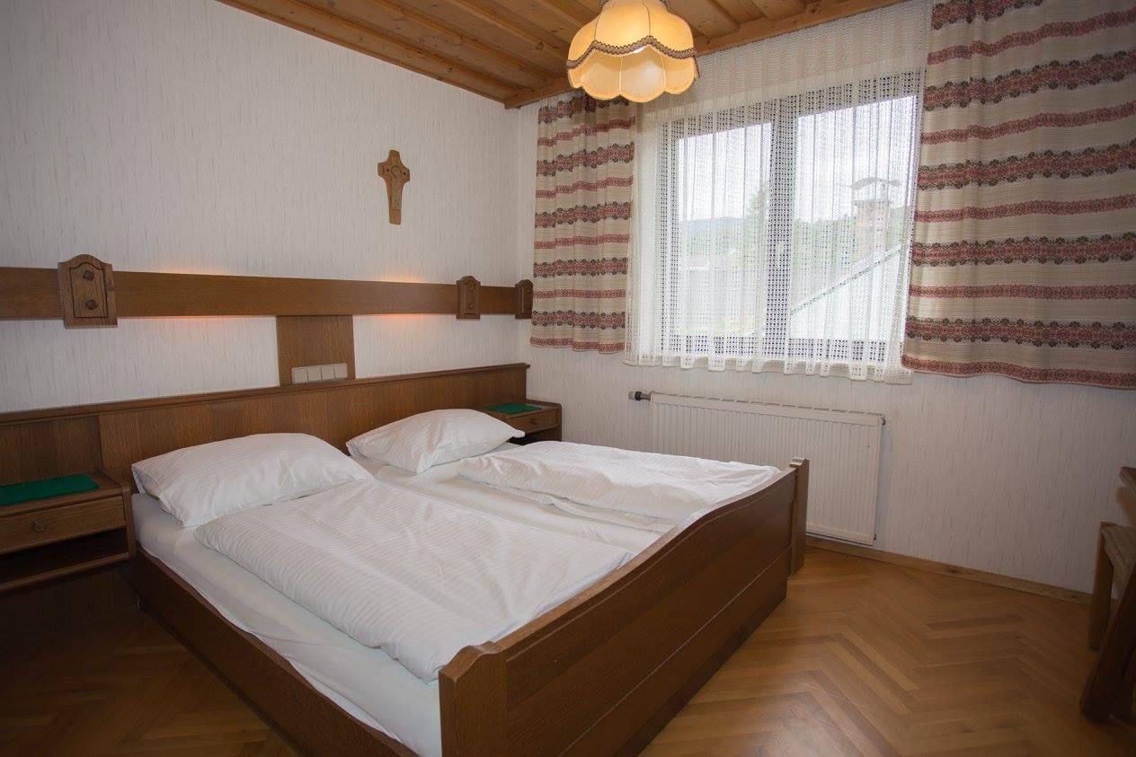 A traditionally furnished double room with wooden furniture, a double bed, a cross on the wall and a window with curtains.