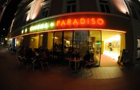 Exterior view of a movie theater called Cinema Paradiso at night with illuminated entrance and people at tables.