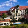 A hotel complex with red roofs, pool and garden, surrounded by trees and flowering plants under a blue sky.