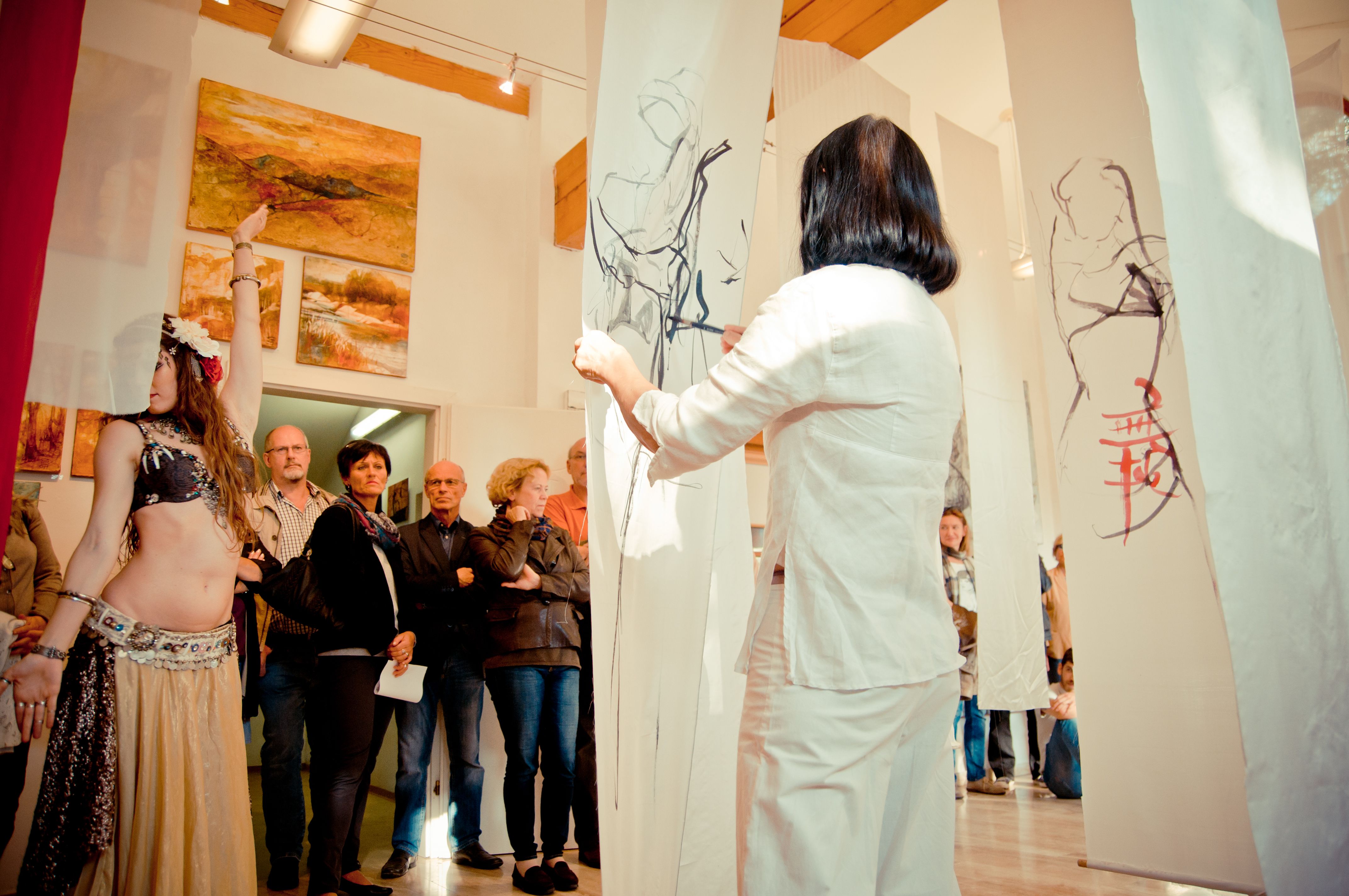 A woman dressed in white looks at drawings on hanging fabric panels in a gallery with spectators and paintings on the wall.