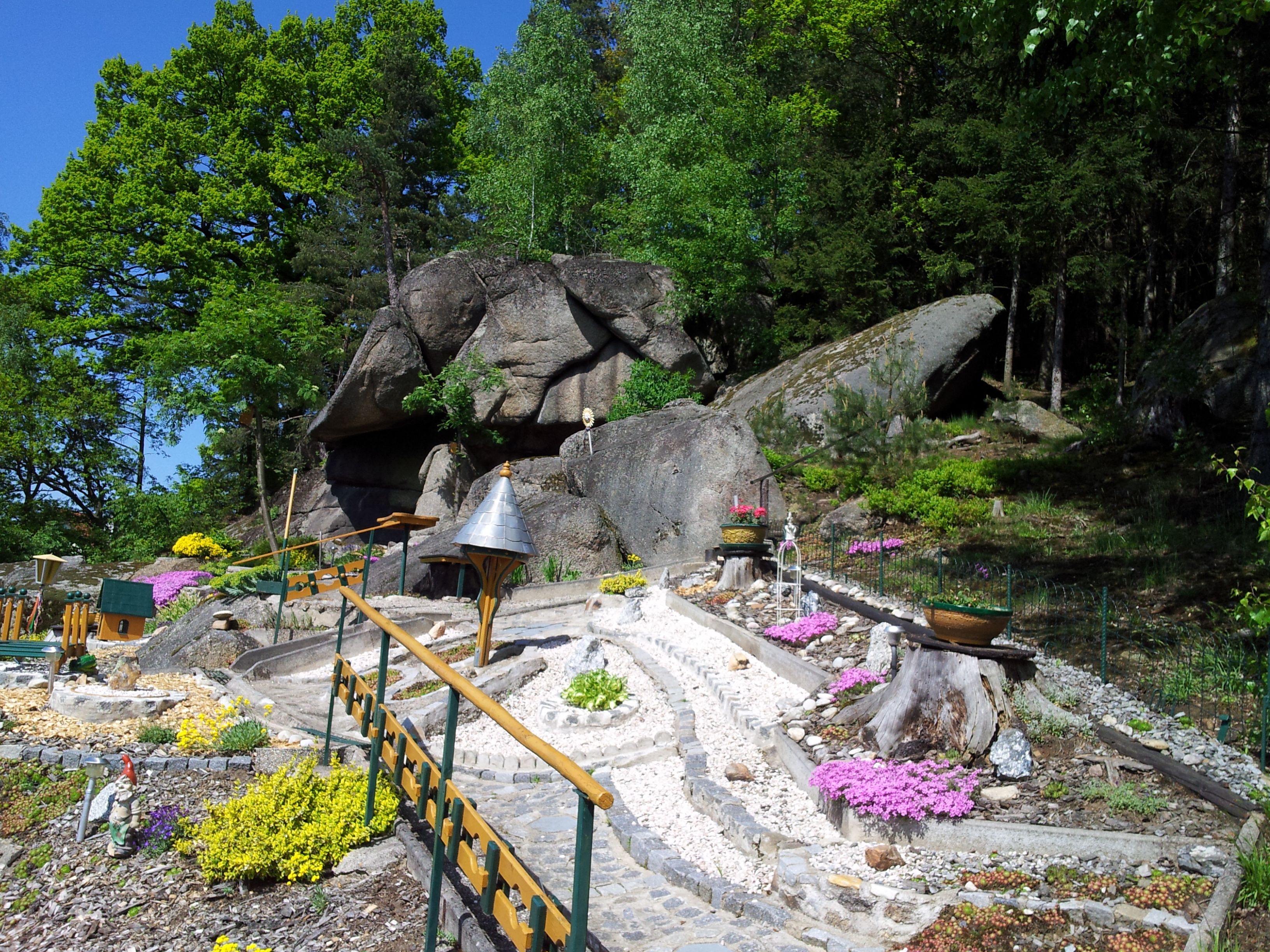 A well-kept garden with stones, flowers and a small path in front of large rocks and trees.