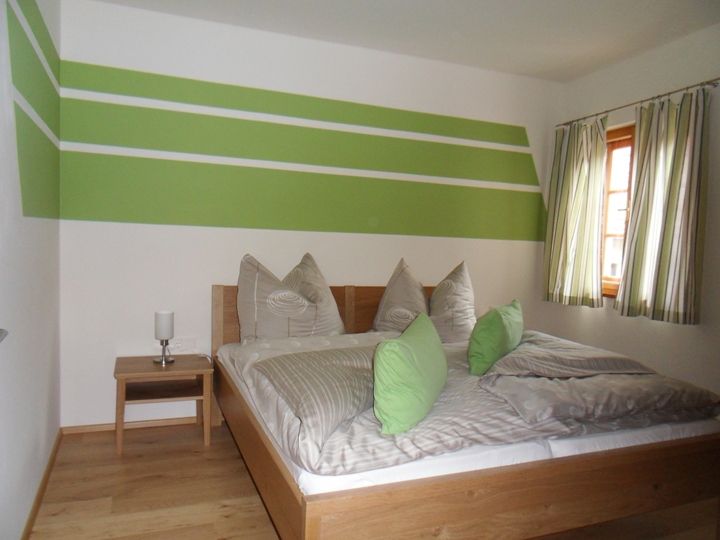 Bedroom with green and white wall decoration, double bed with green cushions, wooden floor and window with striped curtains.