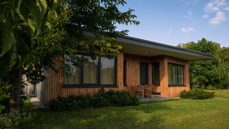 A modern wooden house with large windows, surrounded by trees and lawn.