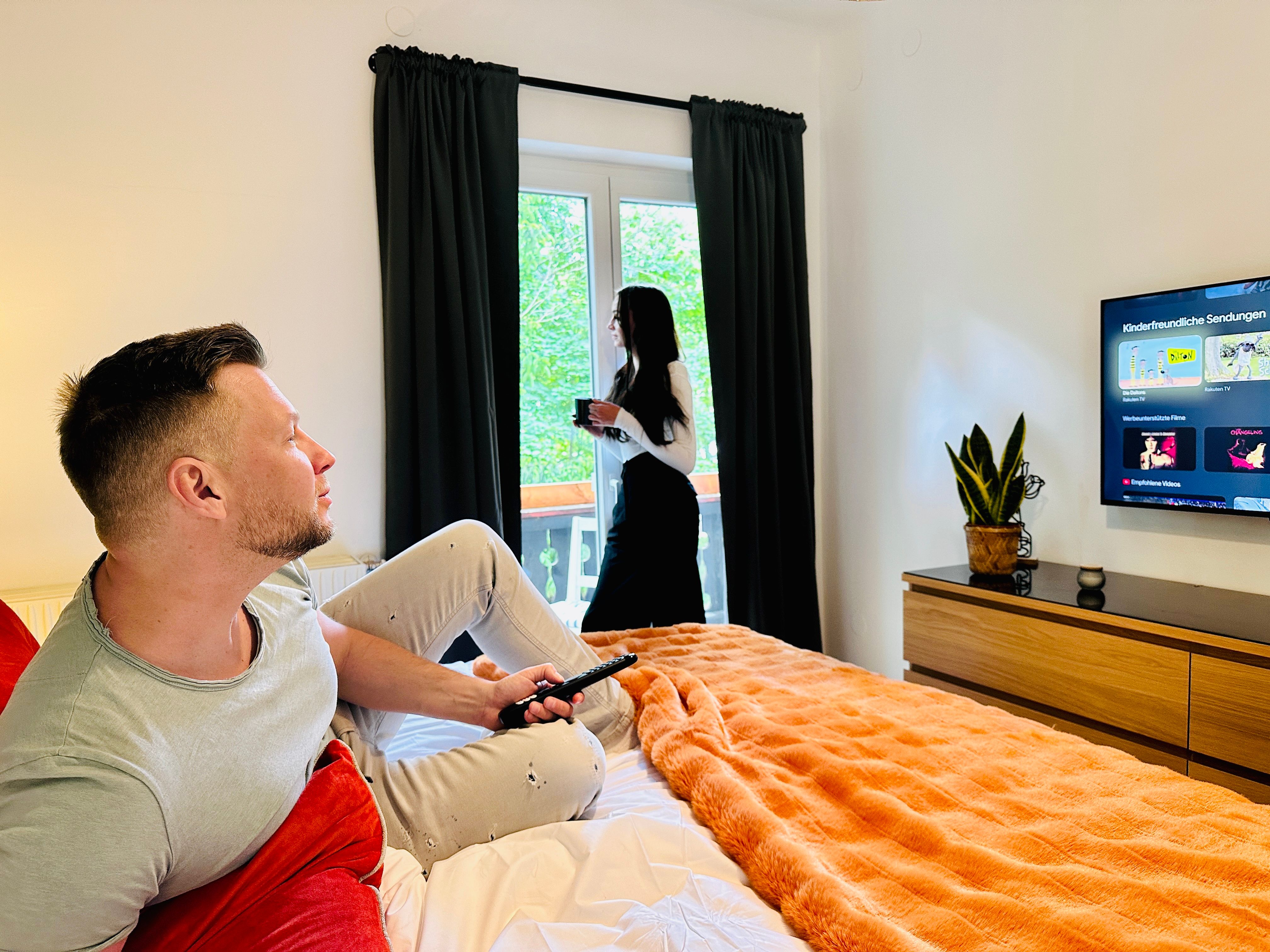 A man sits on a bed and watches television while a woman stands at the window drinking coffee.