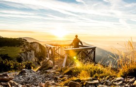 Person stands on a wooden platform and gazes into the sunset over a mountain landscape.