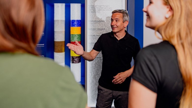 A man explains something to two women in an exhibition room with colorful glass tubes.