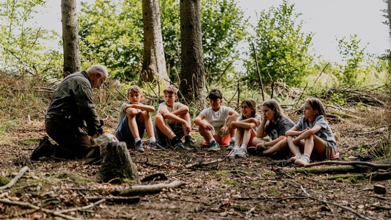 An elderly man speaks to a group of children in the forest.