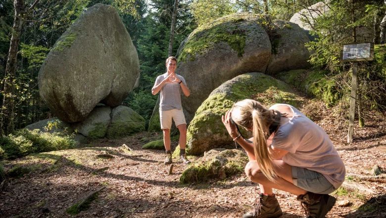 A man poses in front of a large heart-shaped rock in the forest while a woman takes a photo of him.