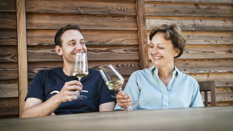 Two people clink glasses of wine, sitting in front of a wooden wall.