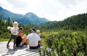 Family sitting on a wooden platform in the Leckermoos high moor, surrounded by mountains and forest.