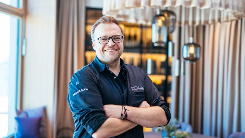 A smiling man in chef's clothes stands with his arms folded in a stylish restaurant.