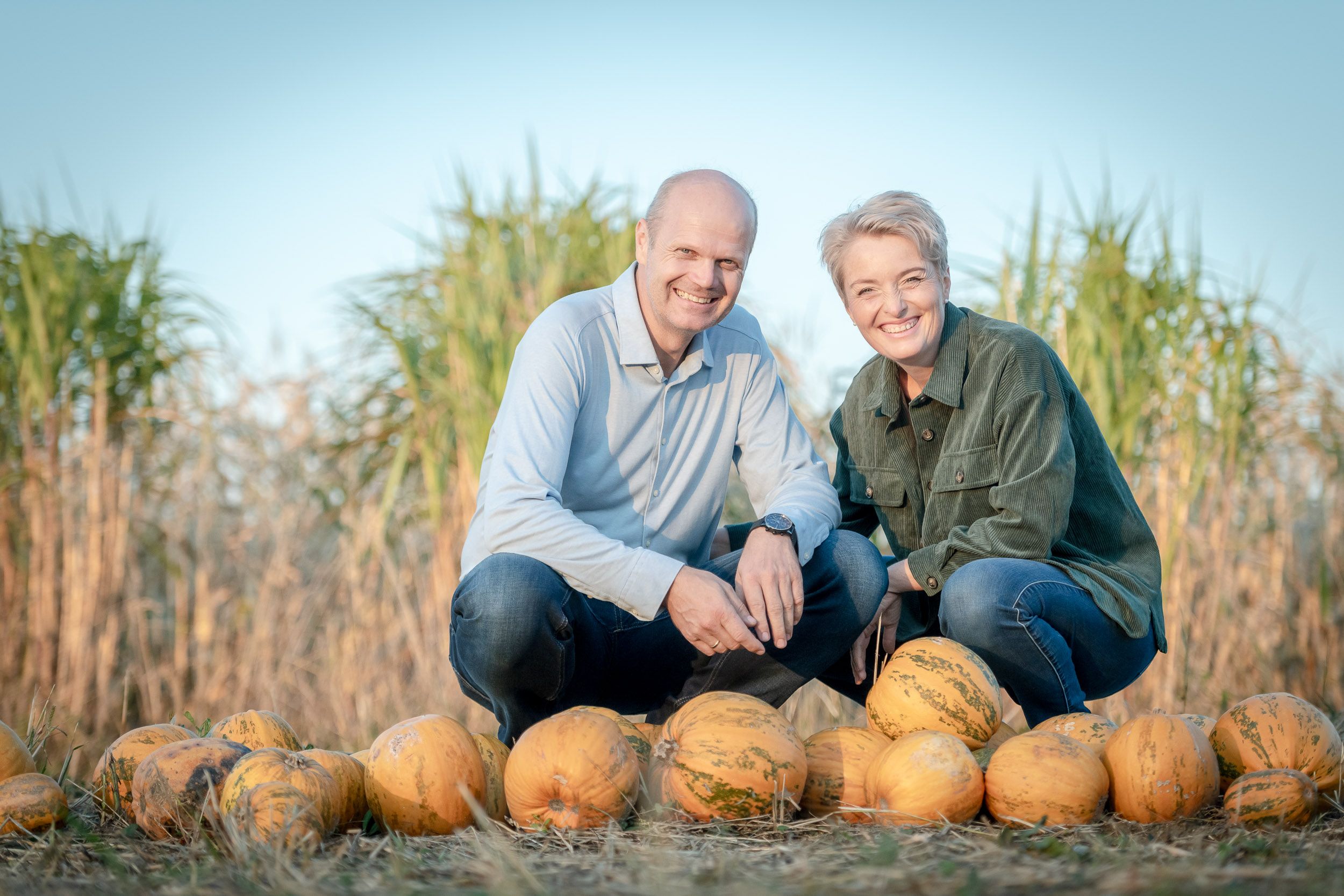 Two people kneel smiling behind a row of pumpkins in a field.