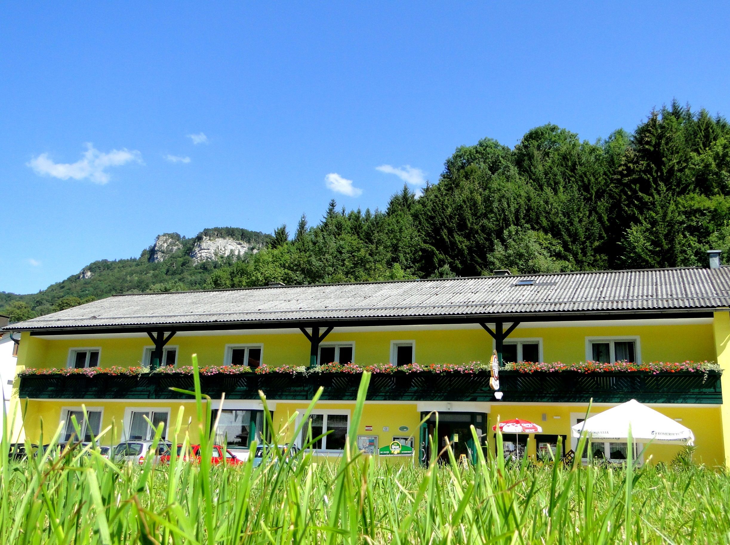Yellow building with flower balcony in front of a wooded hill and blue sky.