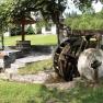 Old mill wheel with millstone, next to steps and a fountain, surrounded by meadow and trees.