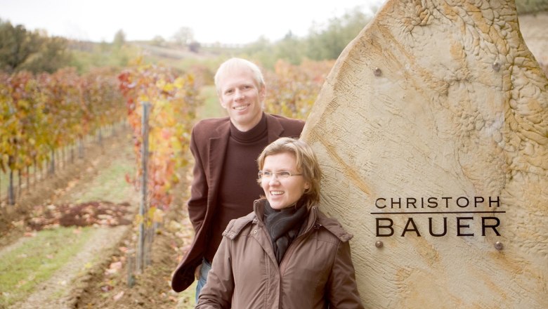 Two people stand smiling in front of a vineyard next to a stone with the inscription 'Christoph Bauer'.