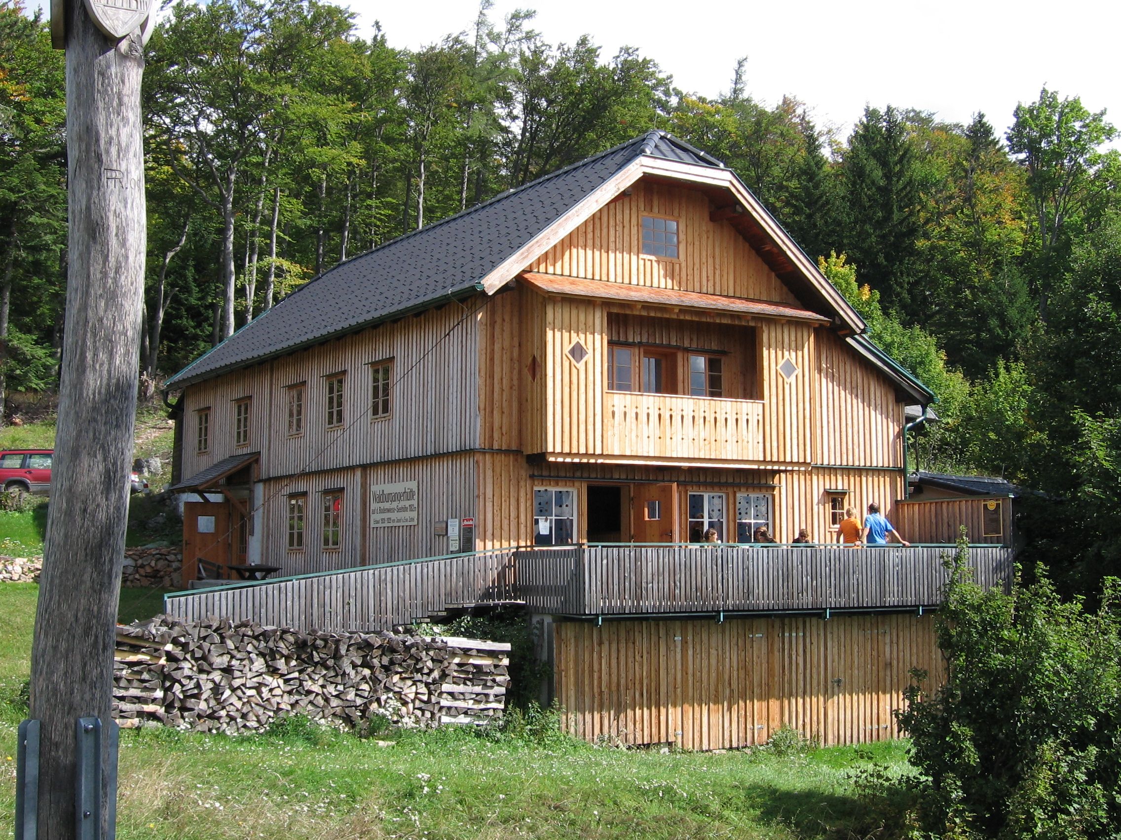 A wooden hut in the forest with a terrace and people on it.