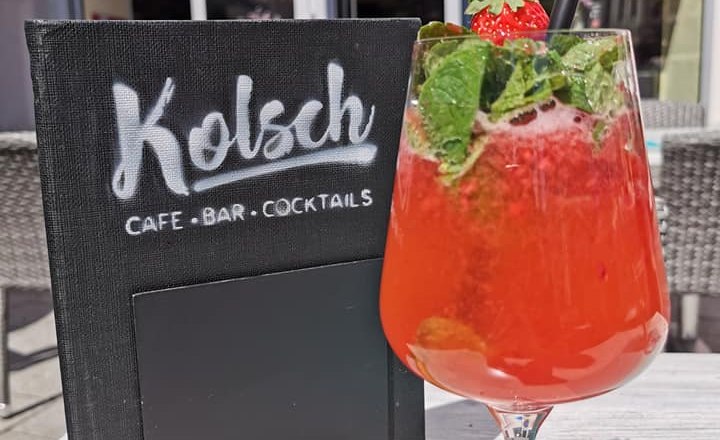 A red cocktail with mint and strawberry in front of a café with the sign 'Kölsch Café Bar Cocktails'.