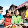 Three people sit laughing at a table in front of a hut while a man in a yellow shirt serves drinks.