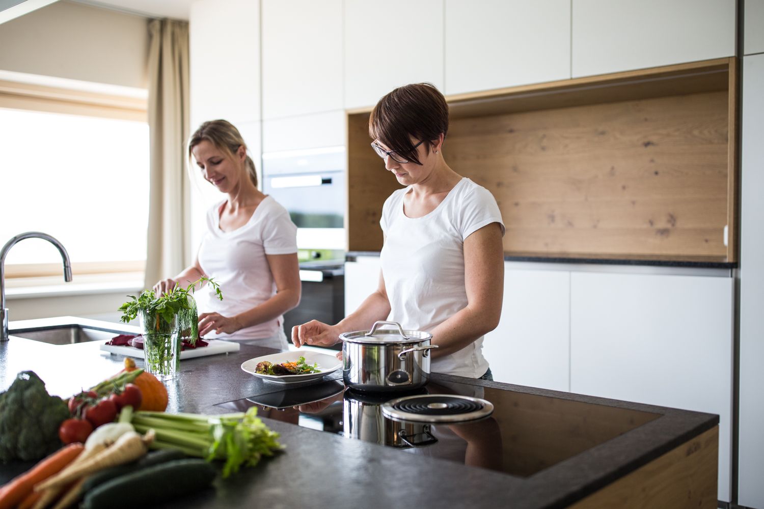Two women cooking in a modern kitchen with fresh vegetables on the worktop.