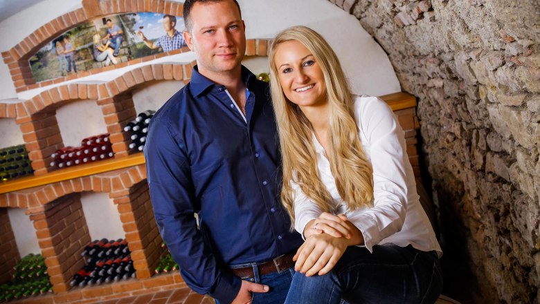 A man and a woman pose in front of wine racks in a wine cellar.