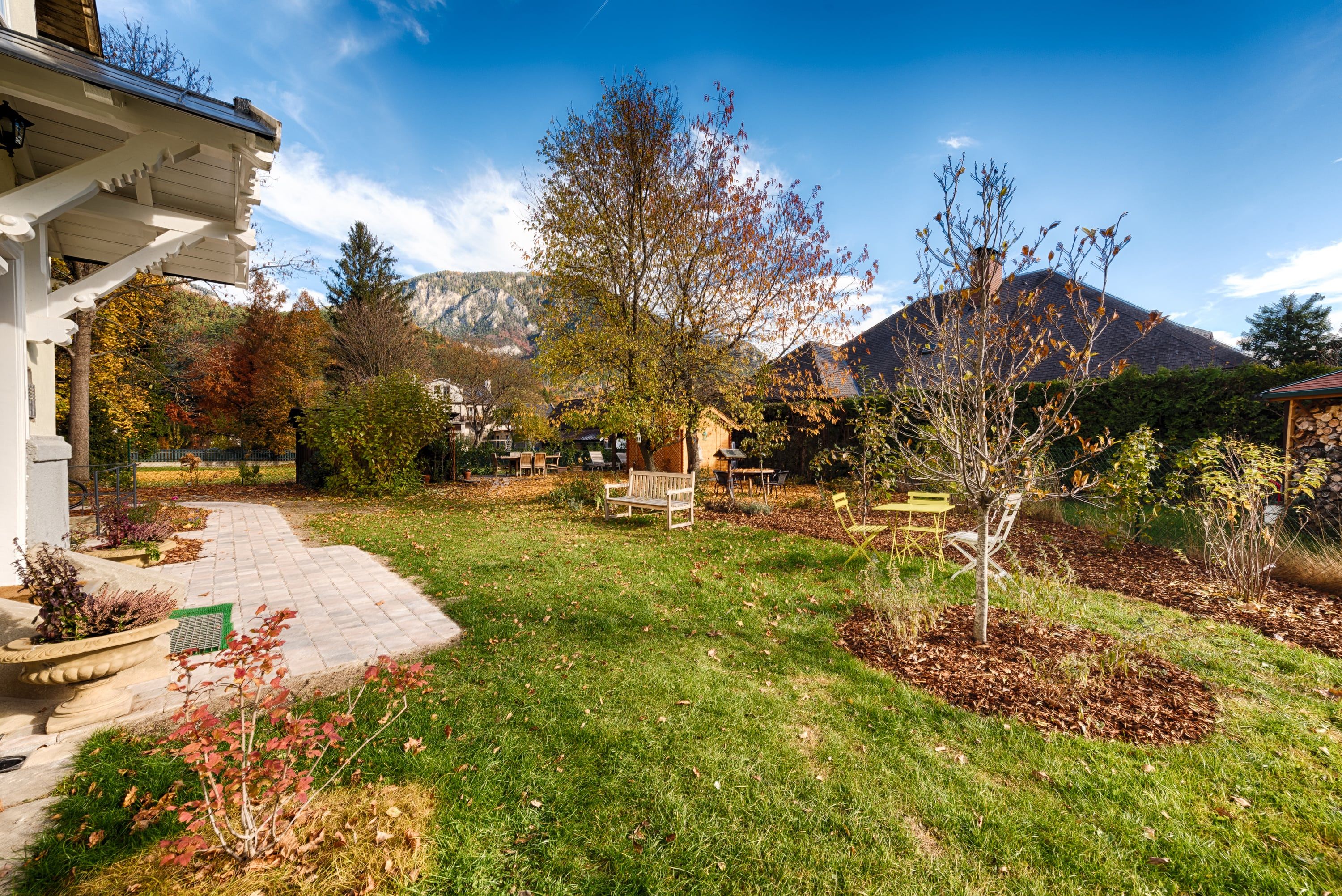 An autumnal garden with trees, benches and a paved path. Mountains and a blue sky can be seen in the background.