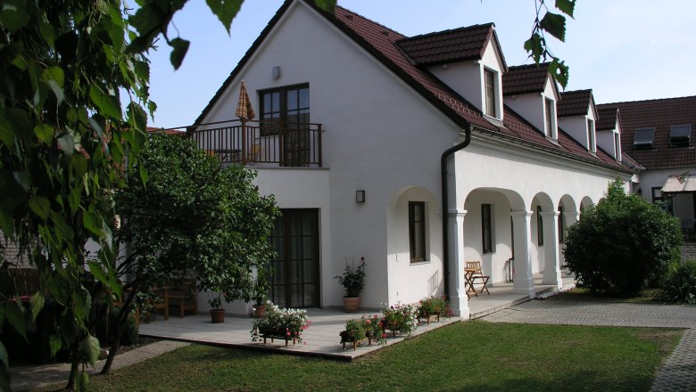 A white building with red roof tiles and arcades, surrounded by a garden with trees and flowers.