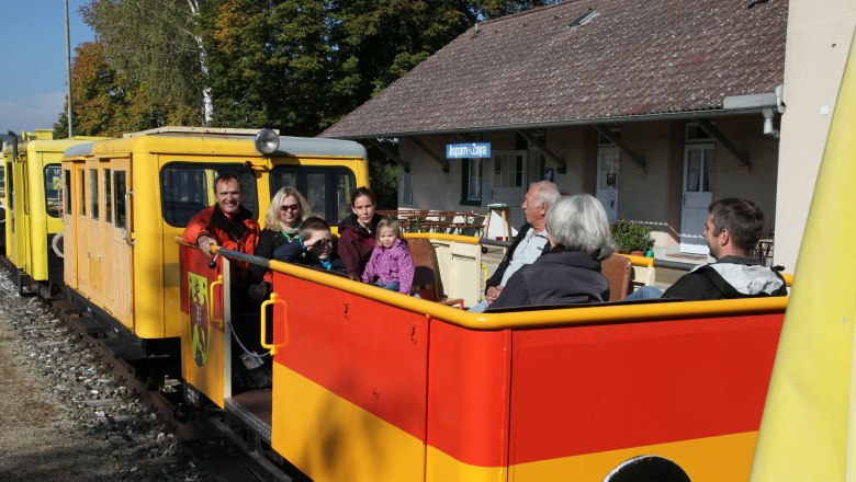 People sit in a yellow and red rail cab in front of a station building.