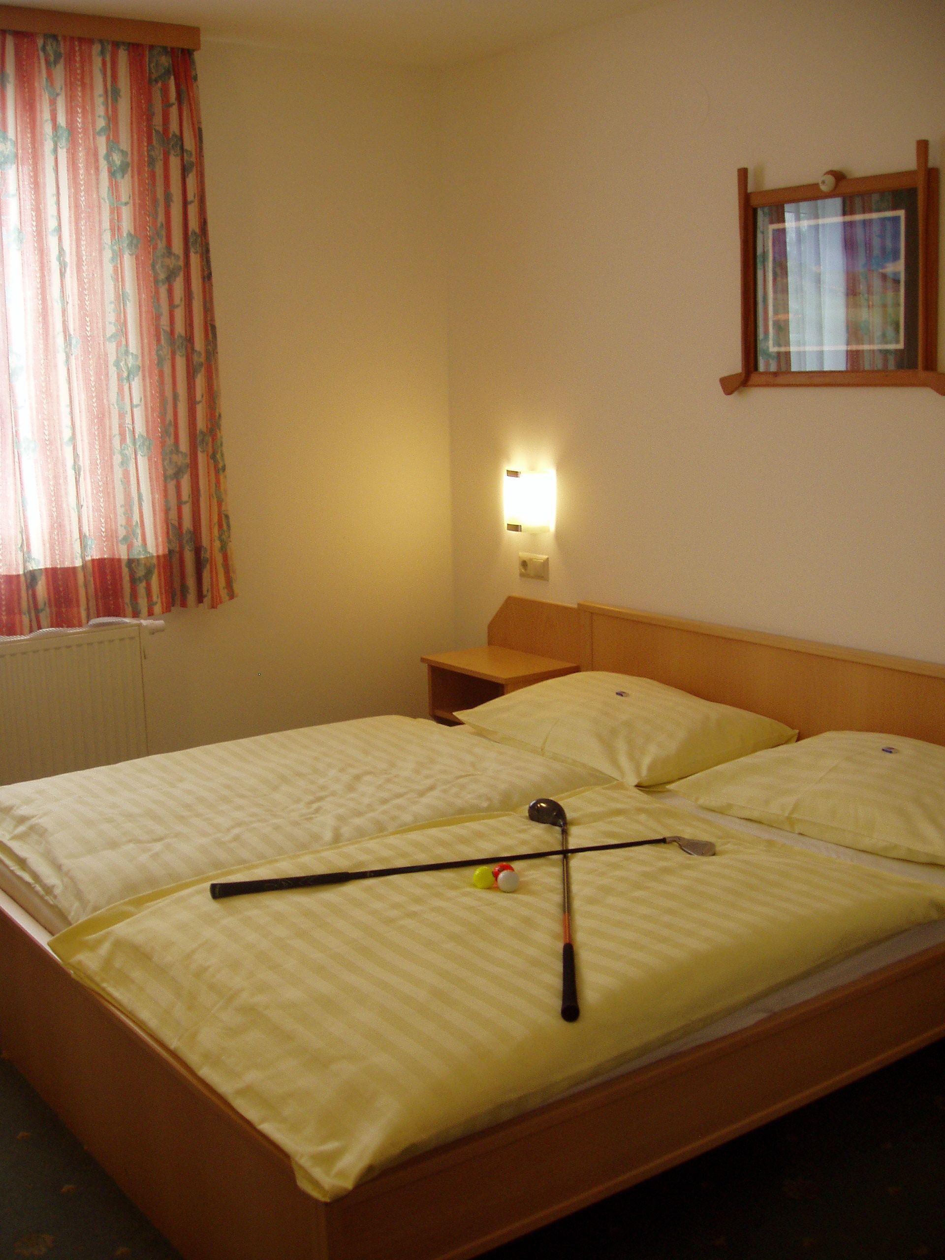 A double bed with yellow bed linen, on which lie two golf clubs and a golf ball. A window with a curtain and a picture on the wall.