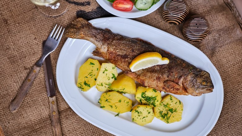 A plate with fried trout, potatoes and a slice of lemon, next to a salad and a glass of white wine.