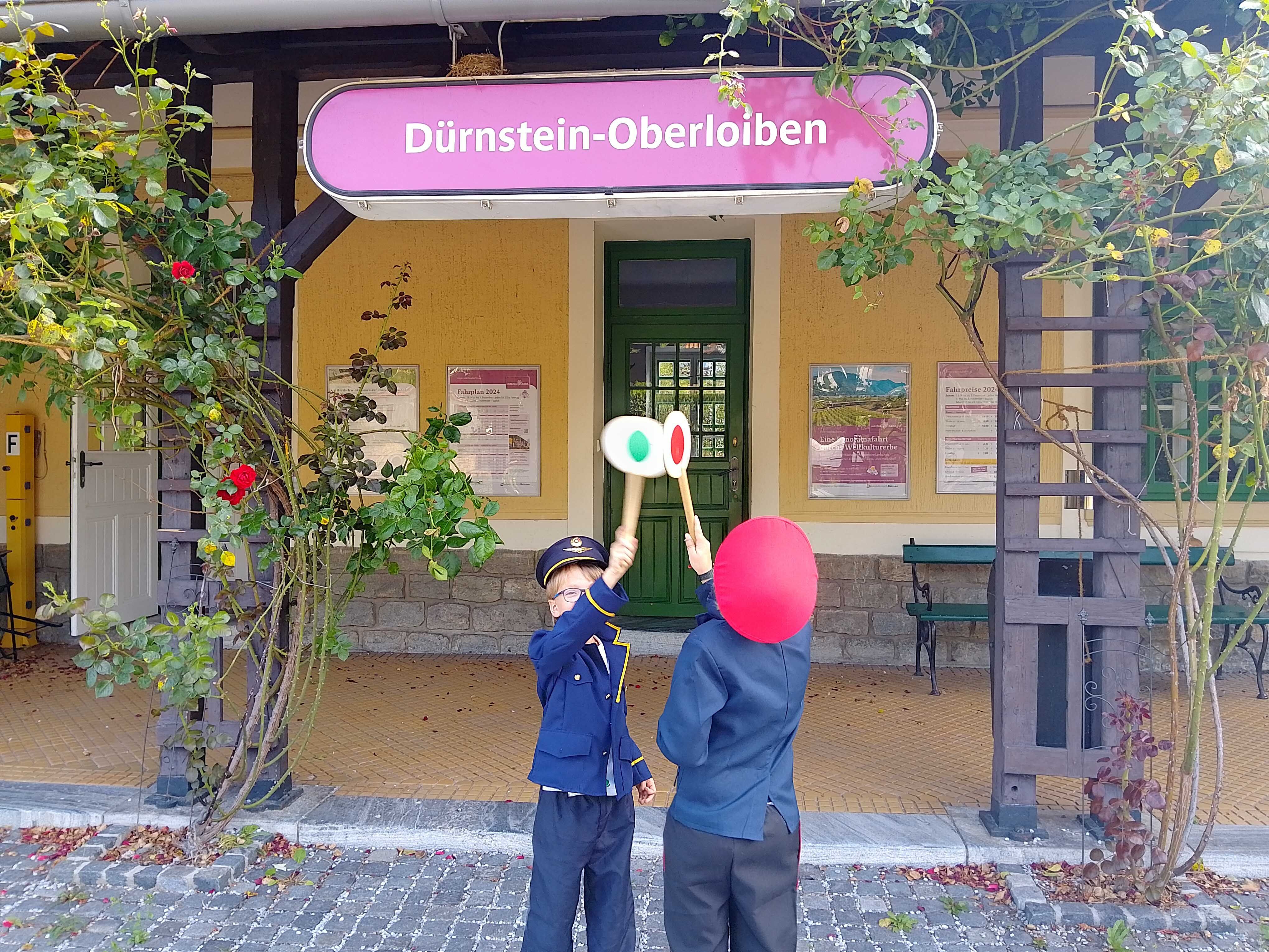 Two children in railroad uniforms stand in front of Dürnstein-Oberloiben station holding up signal boards.