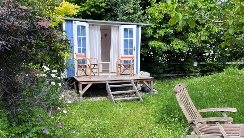 A blue garden pavilion with two chairs on a wooden veranda, surrounded by a green meadow and colorful flowers.