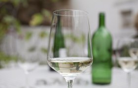 A wine glass with white wine on a table, a green wine bottle in the background.