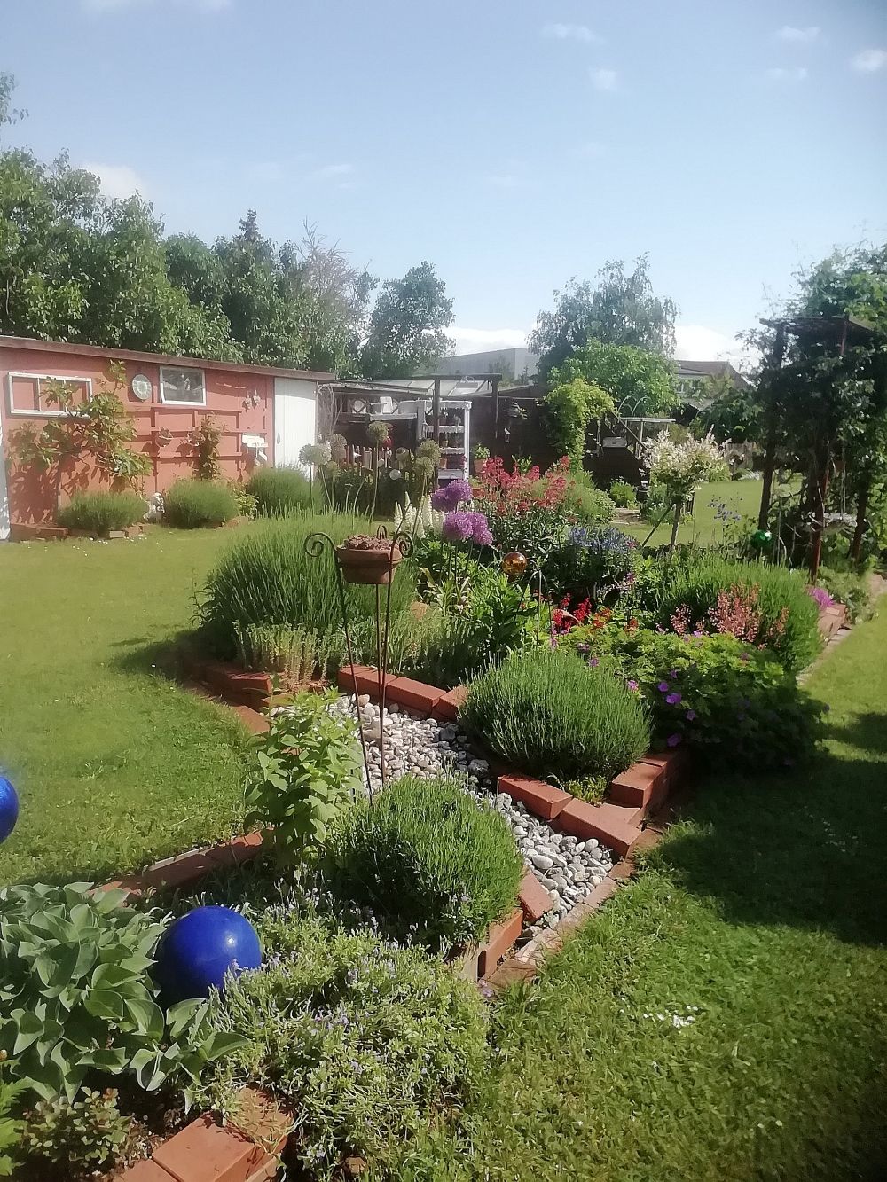 A well-tended garden with shrub beds, brick borders and flowering plants under a blue sky.