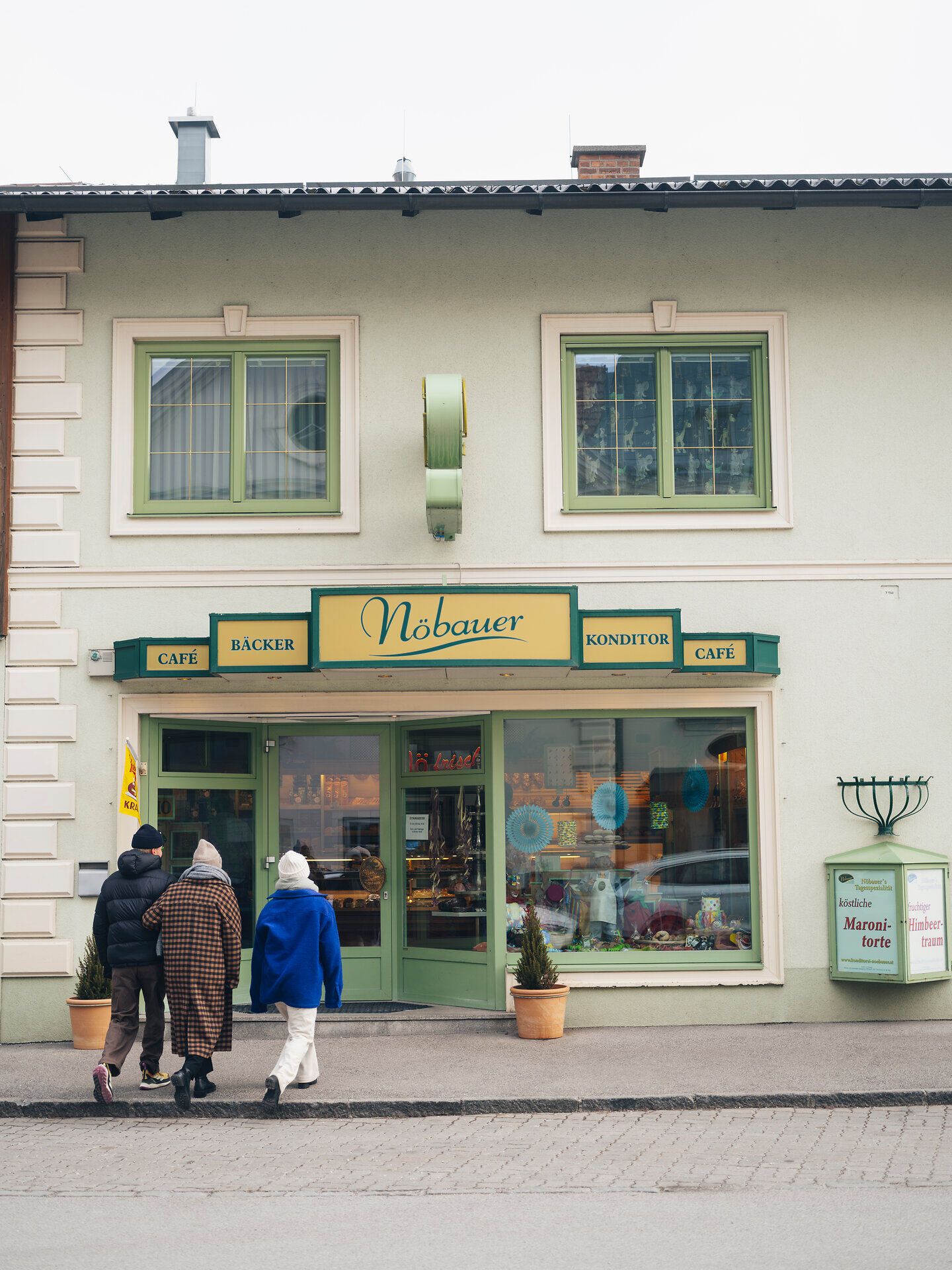 In the wintry atmosphere, visitors stroll relaxed to the charming patisserie with its inviting windows and warm light. The aroma of freshly baked treats wafts through the cold air and promises sweet delights, while the snow-capped mountains in the background provide a picturesque backdrop.