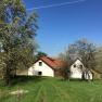 A white house with a red roof stands on a green meadow, surrounded by blossoming trees under a clear blue sky.