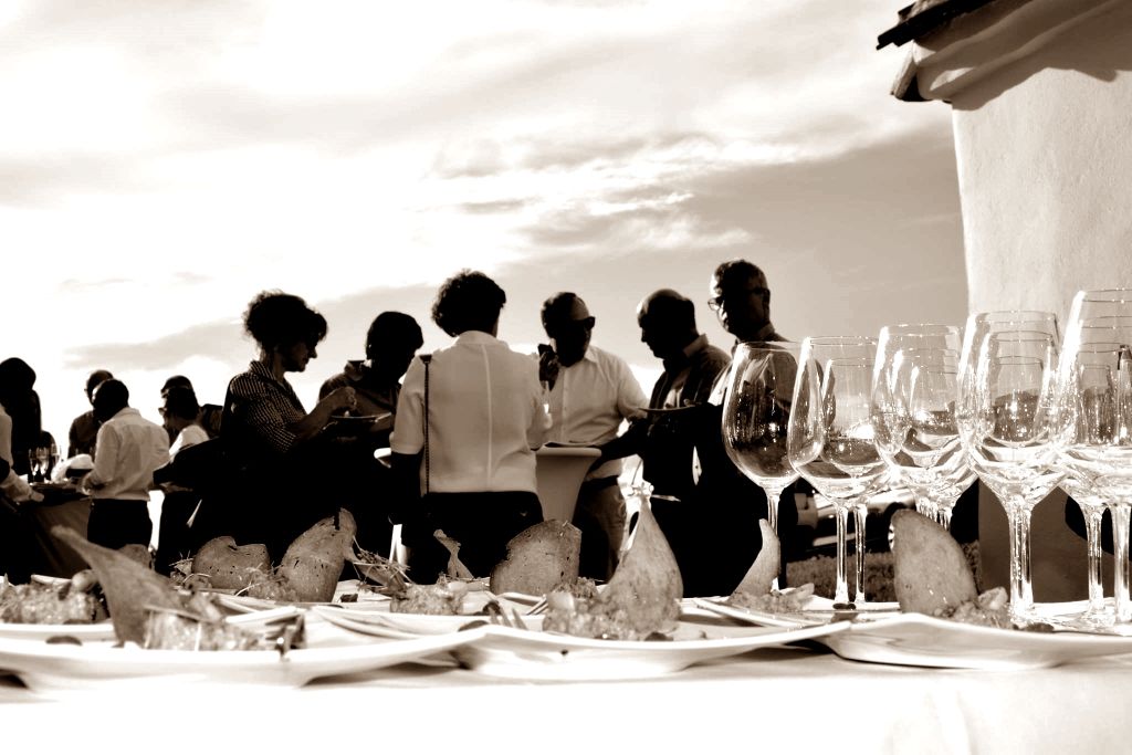 People at an outdoor event with wine glasses and snacks on a table.