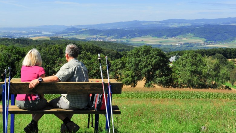 A couple sits on a bench with walking sticks and looks out over a hilly landscape.