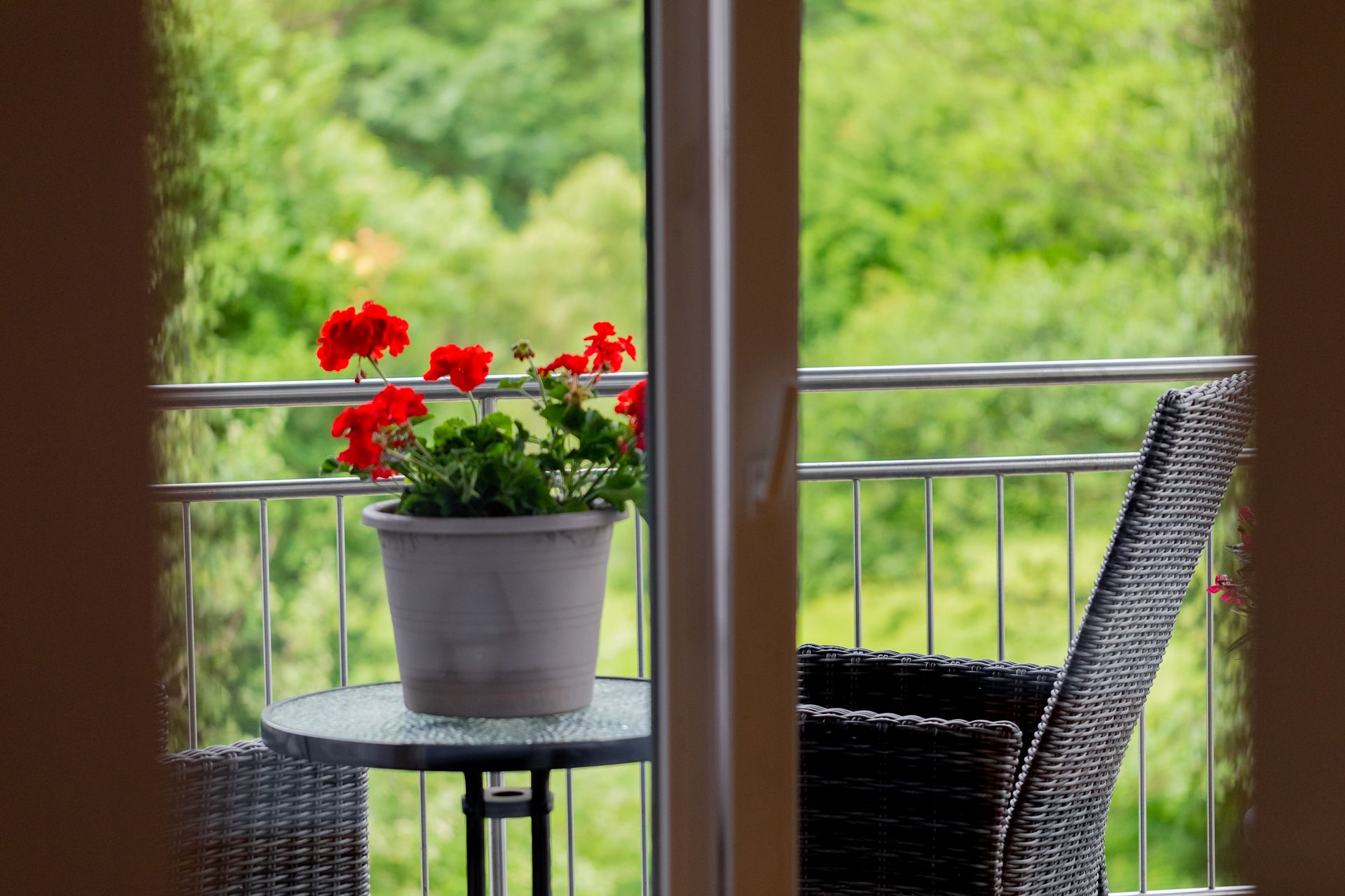 View of a balcony with a table, a flower pot with red flowers and a chair, surrounded by green vegetation.
