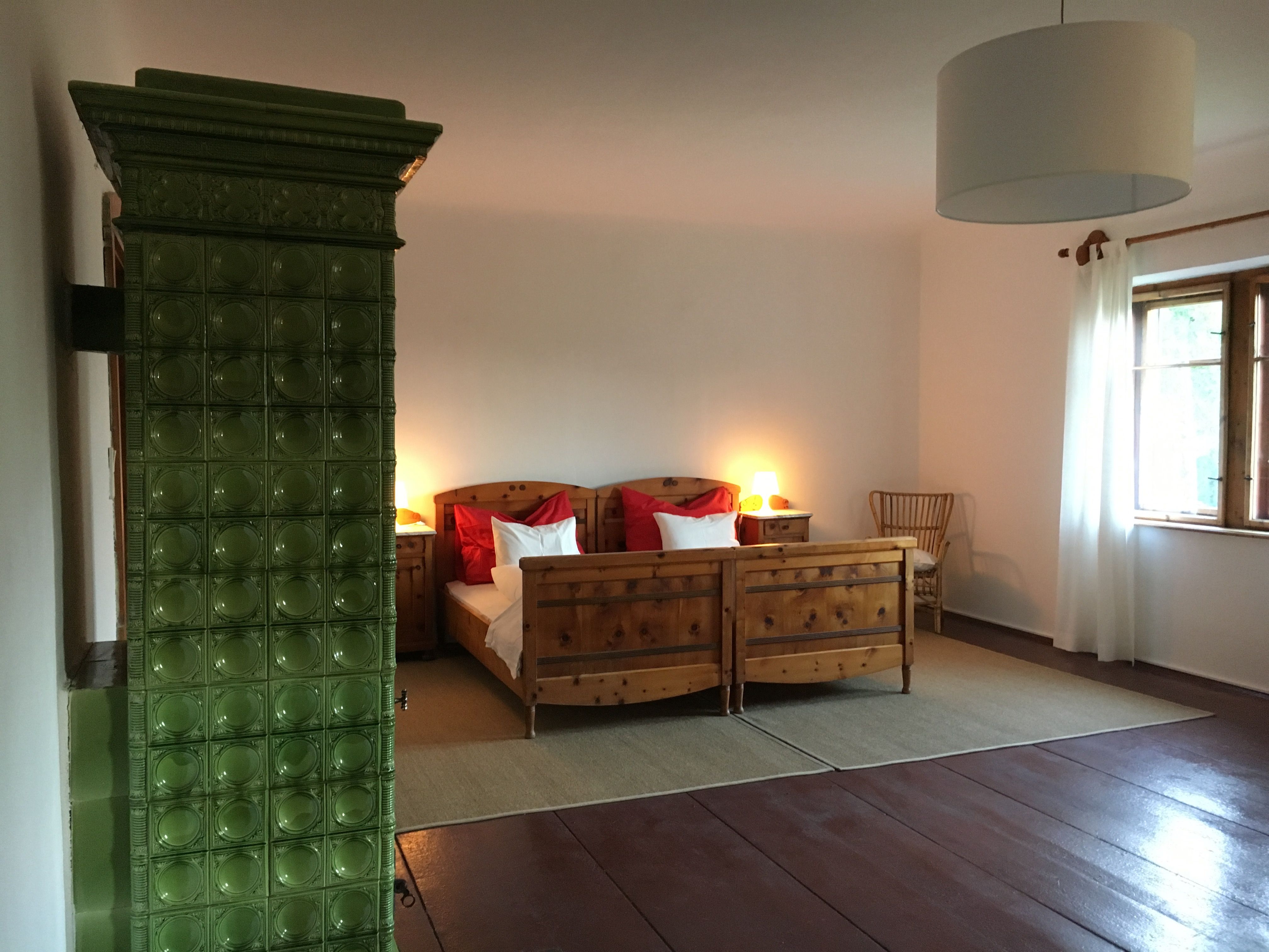 A cozy double room with wooden furniture, a green tiled stove and a large window with white curtains.