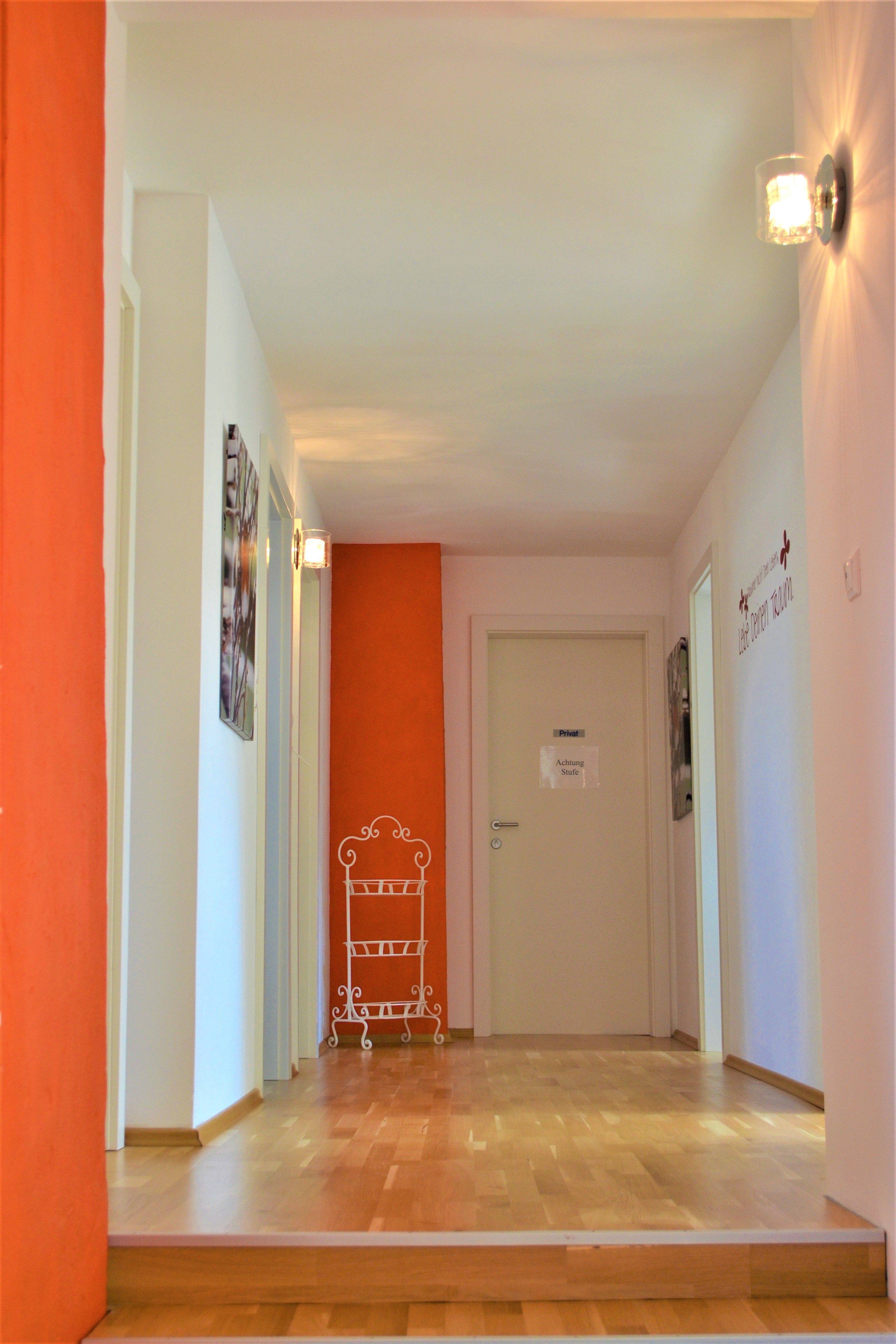 A hallway with a wooden floor, white walls and an orange wall. There is a sign on the wall with the words 'Caution step'.