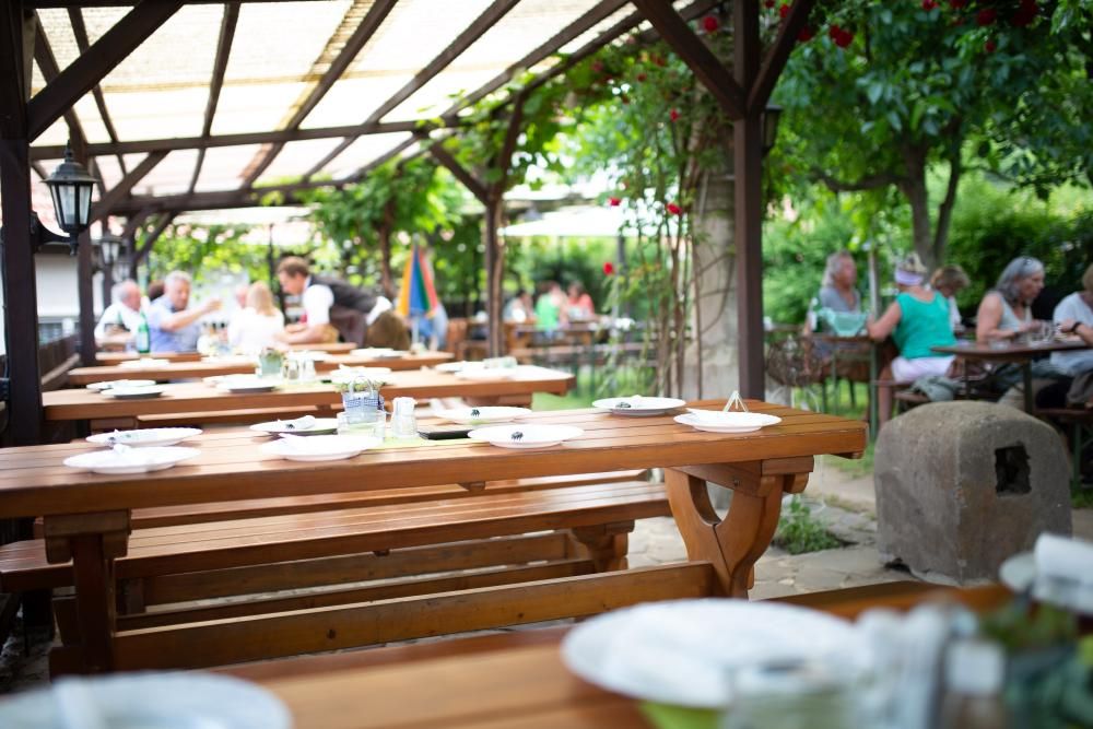 A guest garden with wooden tables and benches, surrounded by green plants. People sit in the background and enjoy their meals.