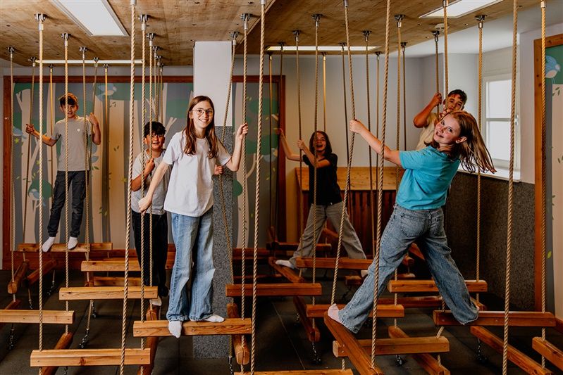 Children play on an indoor ropes course with wooden platforms.