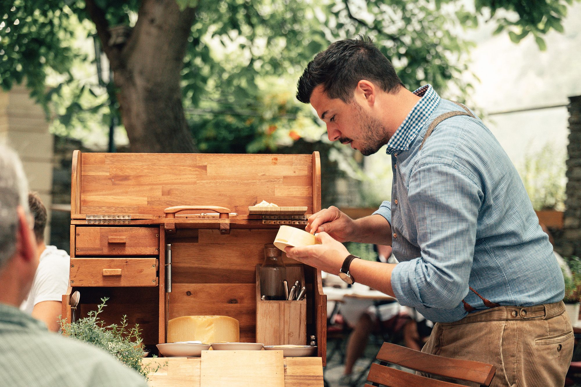 A man in a blue shirt prepares cheese on a wooden serving trolley outside.